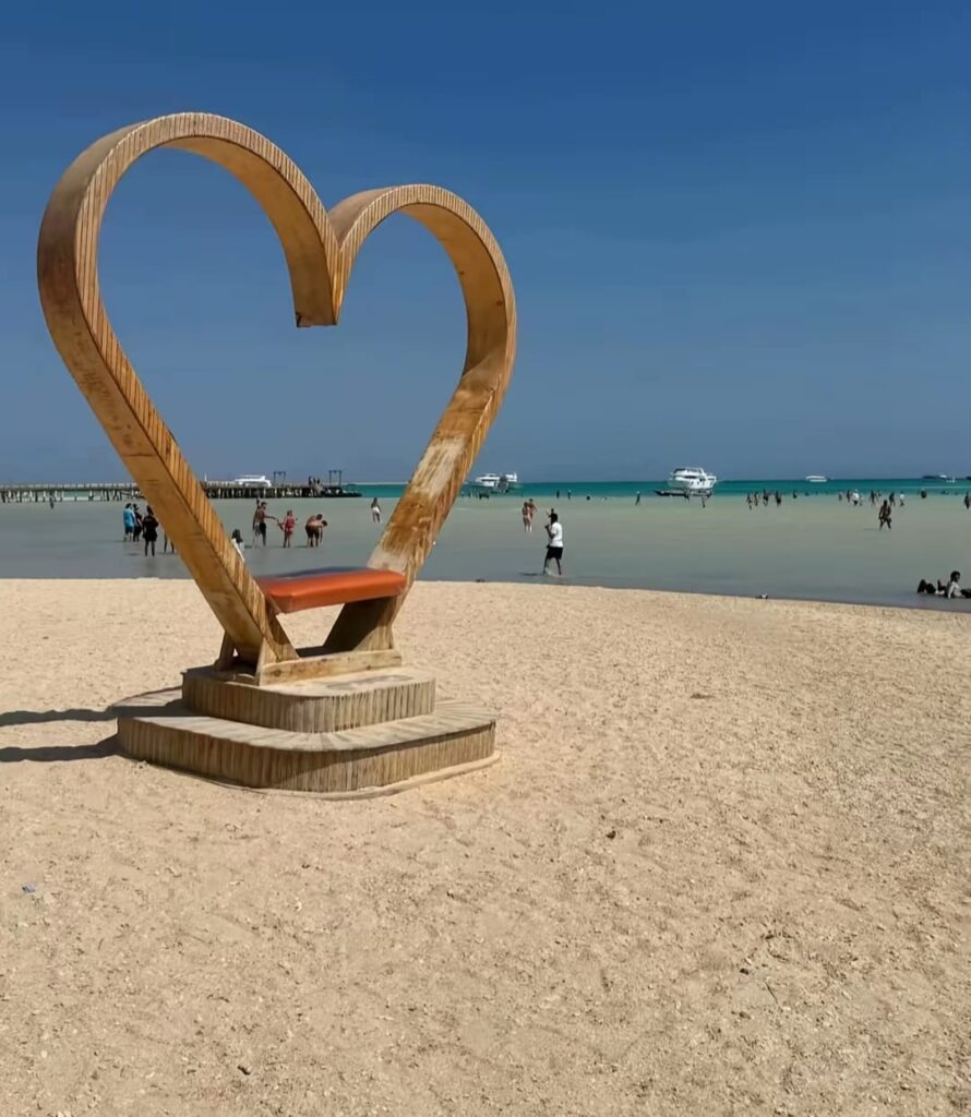 Large wooden heart sculpture on the sandy shore of Orange Bay island with turquoise water and boats